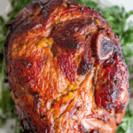 Roasted ham with a caramelized glaze, inspired by a classic glazed ham recipe, surrounded by green garnish, shown close-up on a white plate.