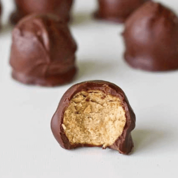 A chocolate-covered round candy with a bite taken out, revealing a creamy peanut butter center. Other whole Cookie Butter Buckeyes are visible in the background.