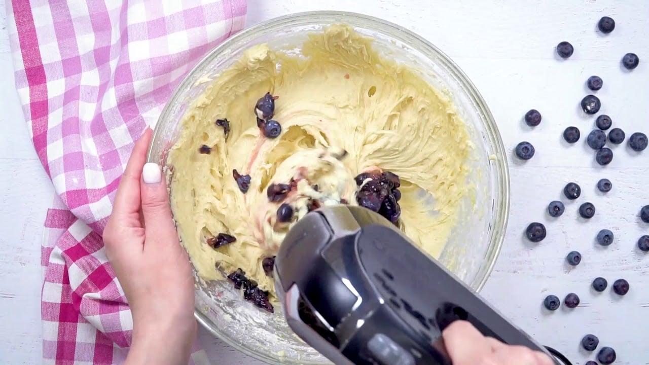 A person using a hand mixer to mix blueberries in a bowl for The Best Blueberry Muffins recipe.