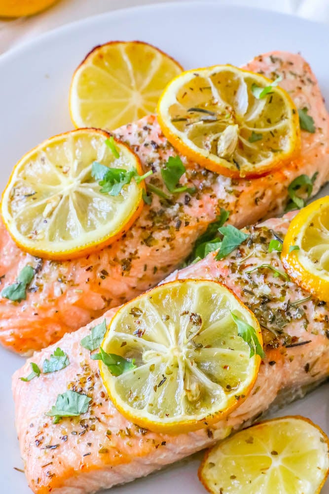 picture of salmon filets with lemon slices, garlic and herbs on top on a white plate
