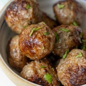 A bowl containing several browned meatballs without breadcrumbs, garnished with chopped parsley.