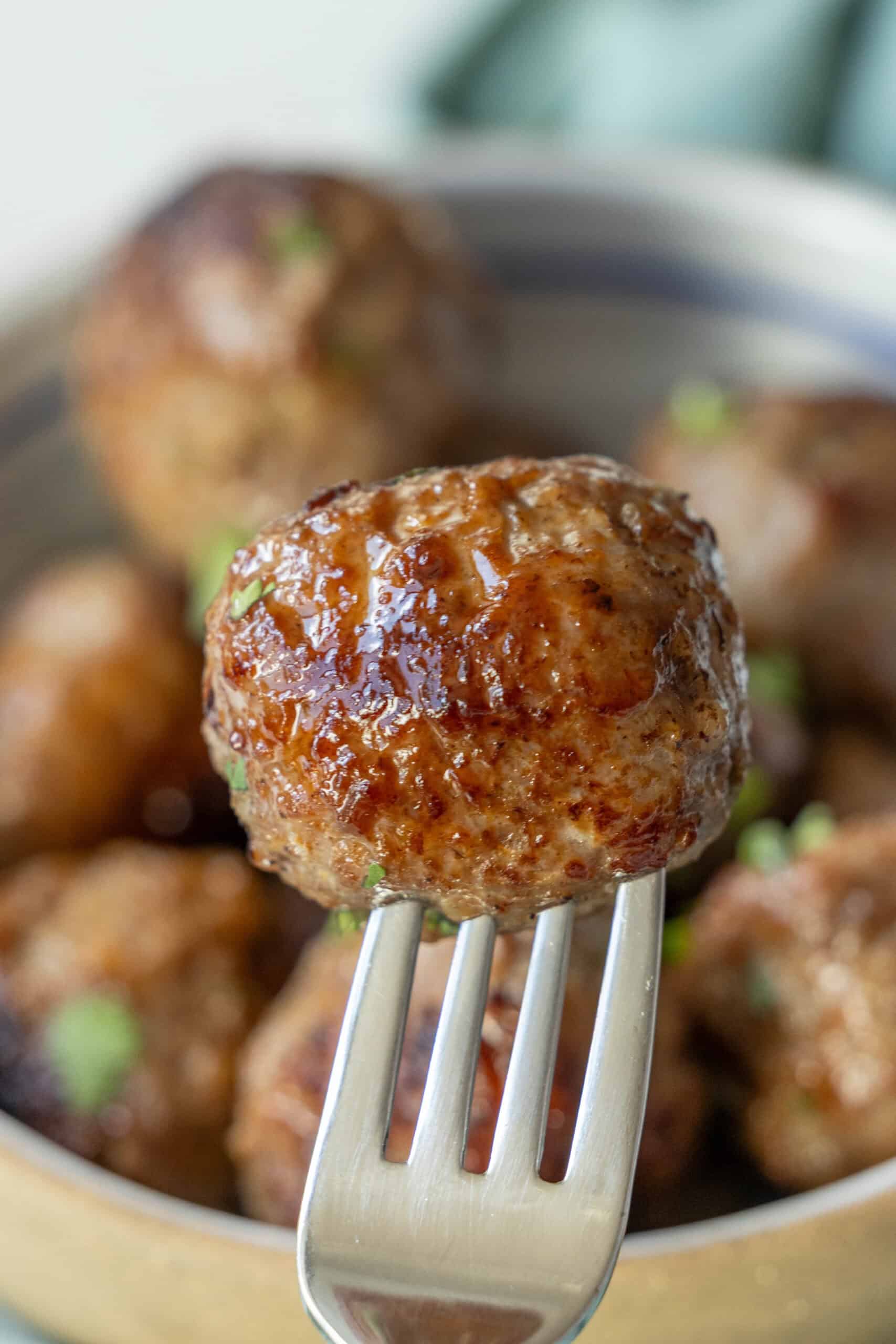 Close-up of a fork holding a perfectly browned, glazed air fried meatball against a backdrop of more meatballs in a bowl.