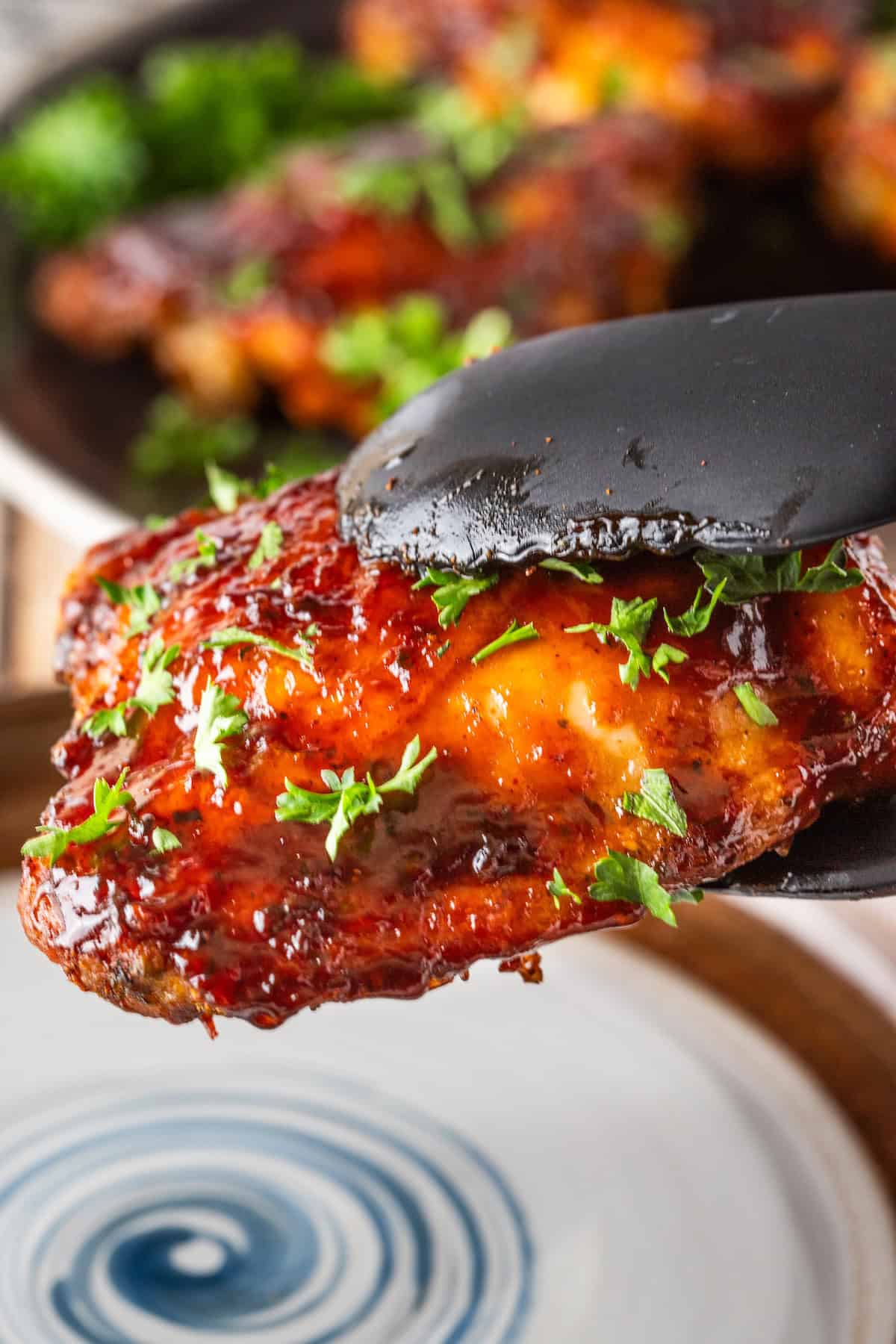 Close-up of black tongs holding a glazed, grilled chicken wing garnished with chopped parsley. Other wings are blurred in the background on a dark plate, reminiscent of those sizzling keto BBQ chicken thighs pan-fried to perfection.