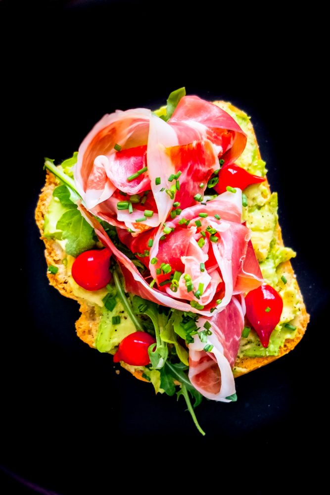 piece of bread with avocado smear, tomatoes, greens, prosciutto, and chives