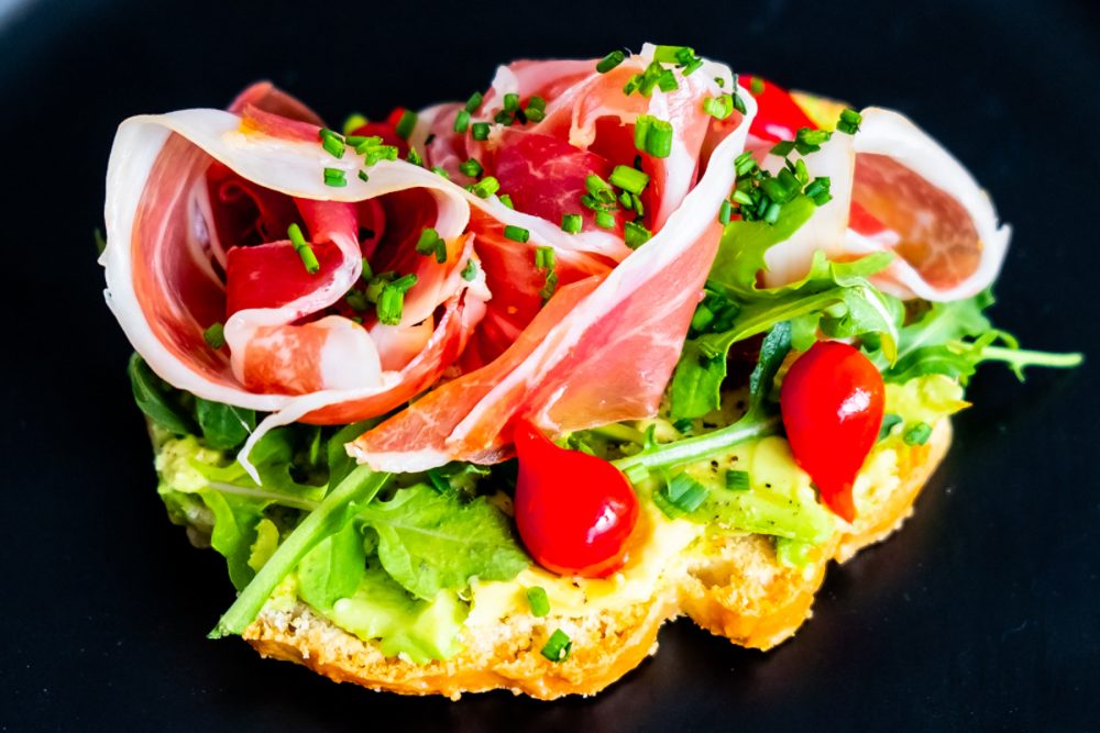 piece of bread with avocado smear, tomatoes, greens, prosciutto, and chives