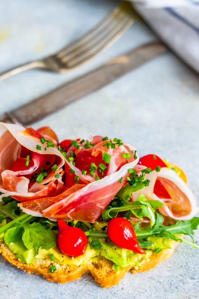 piece of bread with avocado smear, tomatoes, greens, prosciutto, and chives