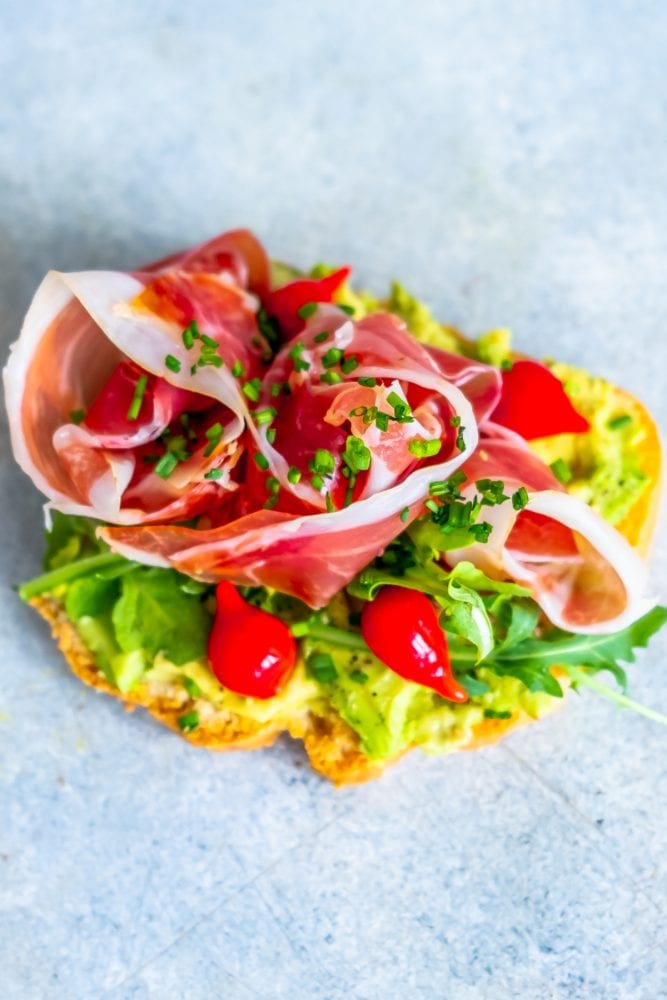 piece of bread with avocado smear, tomatoes, greens, prosciutto, and chives