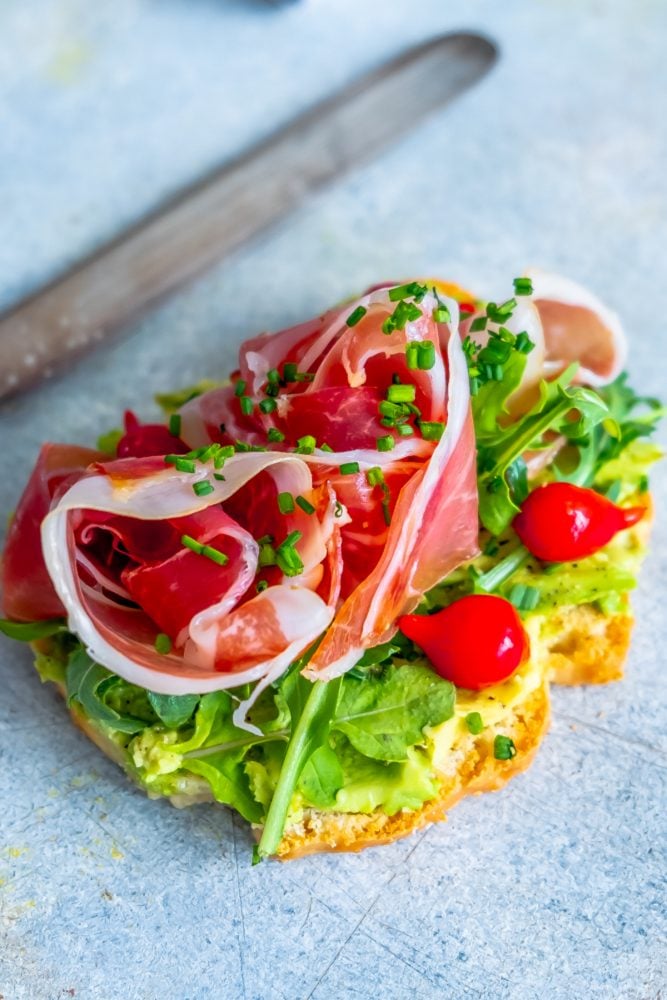 piece of bread with avocado smear, tomatoes, greens, prosciutto, and chives