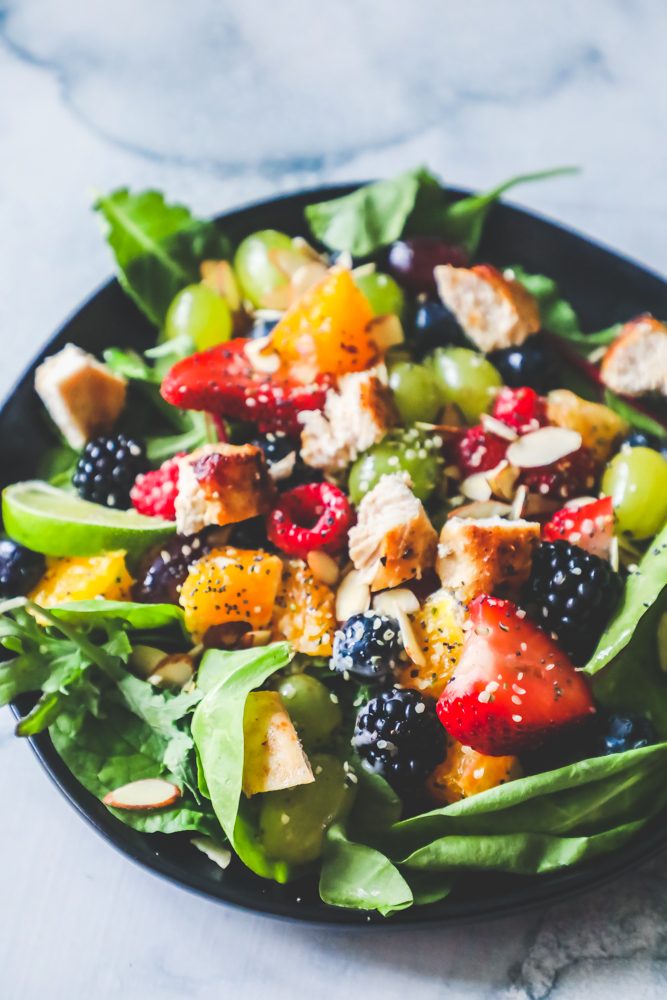 Picture of spinach salad with grapes, strawberries, blackberries, blueberries, raspberries, chicken, poppy seeds, hemp hearts, and a citrus poppyseed dressing on a black plate. 