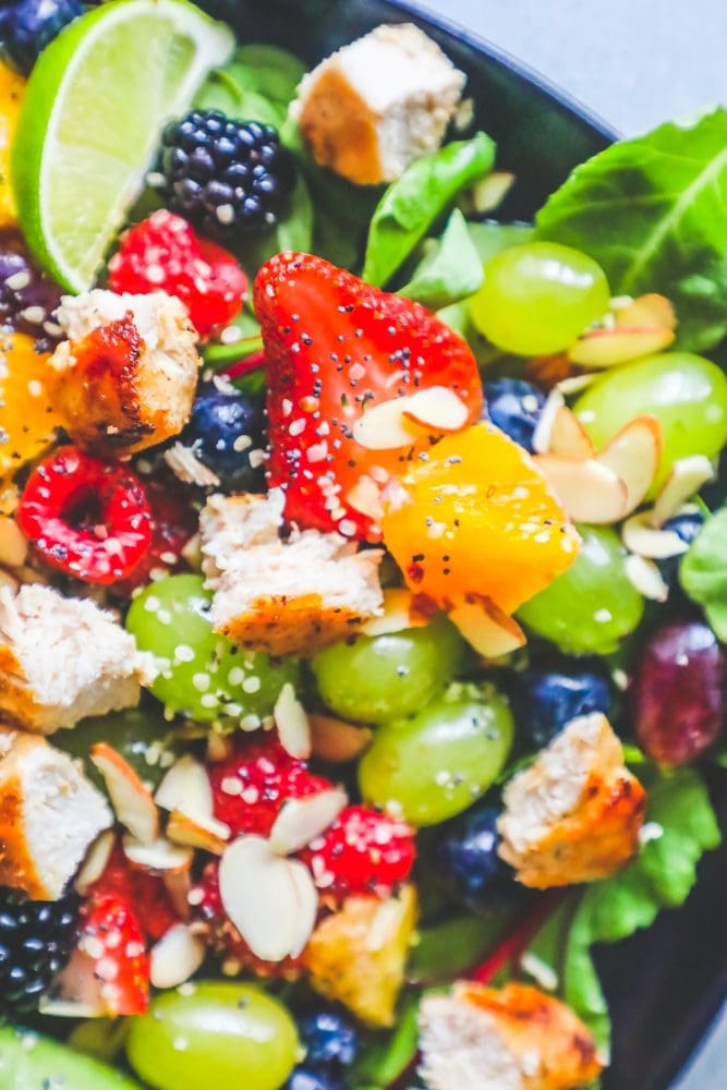Picture of spinach salad with grapes, strawberries, blackberries, blueberries, raspberries, chicken, poppy seeds, hemp hearts, and a citrus poppyseed dressing on a black plate. 