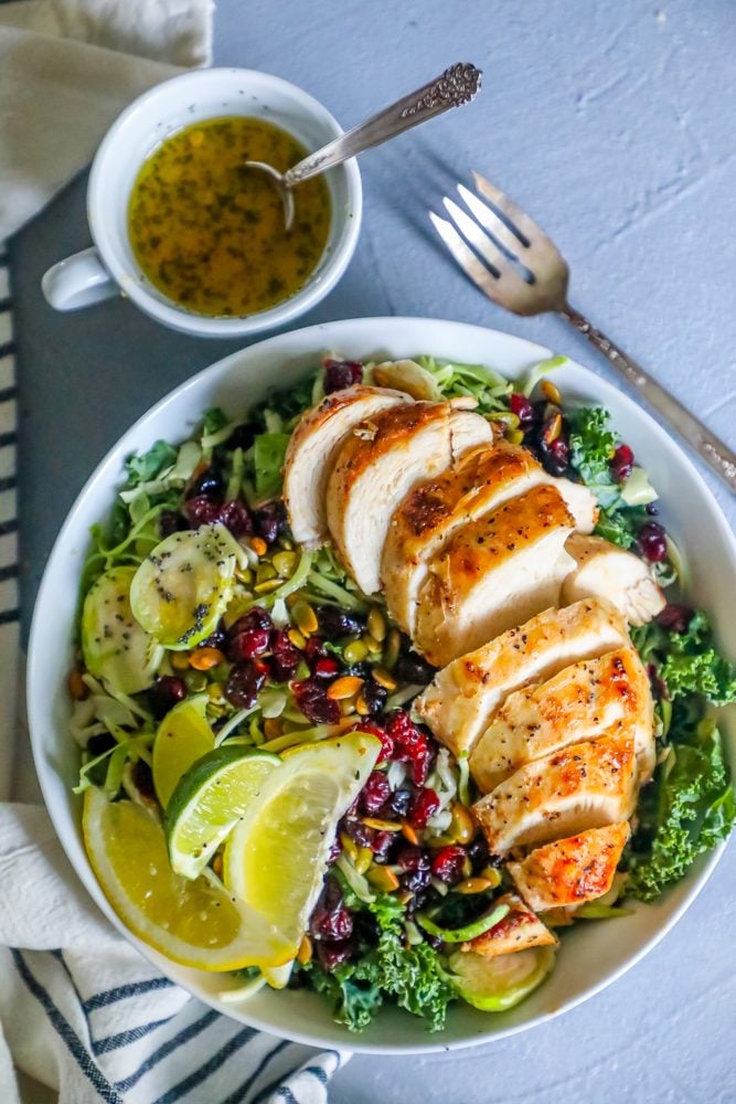 salad with nuts, kale, cranberries, poppyseeds, lemon, lime, and roast chicken in a white bowl