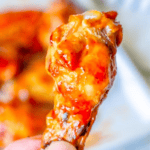 Close-up of a hand holding a chicken wing coated in a thick, reddish-orange sauce from our Baked Crispy BBQ Chicken Wings recipe, with more sauced wings blurred in the background.