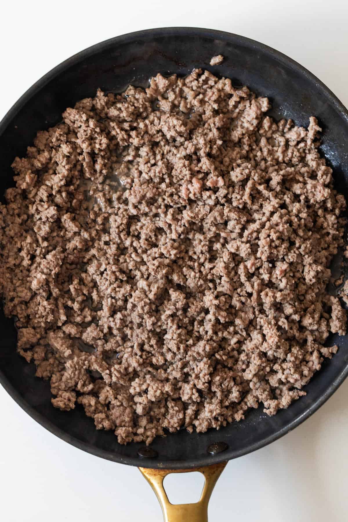 Cooked ground beef in a black skillet with a gold handle, shown from above on a white background—perfect for an easy taco and rice skillet dinner.