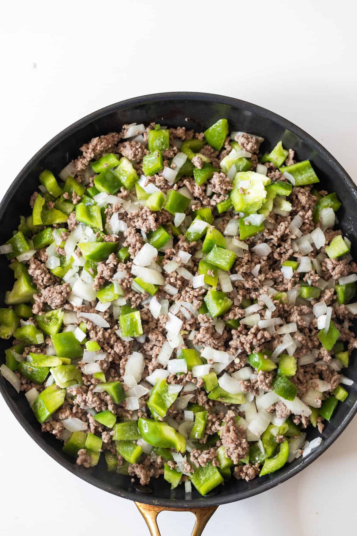 A skillet filled with cooked ground beef, chopped green bell peppers, and diced onions—perfect for an easy taco and rice skillet dinner—set against a white background.