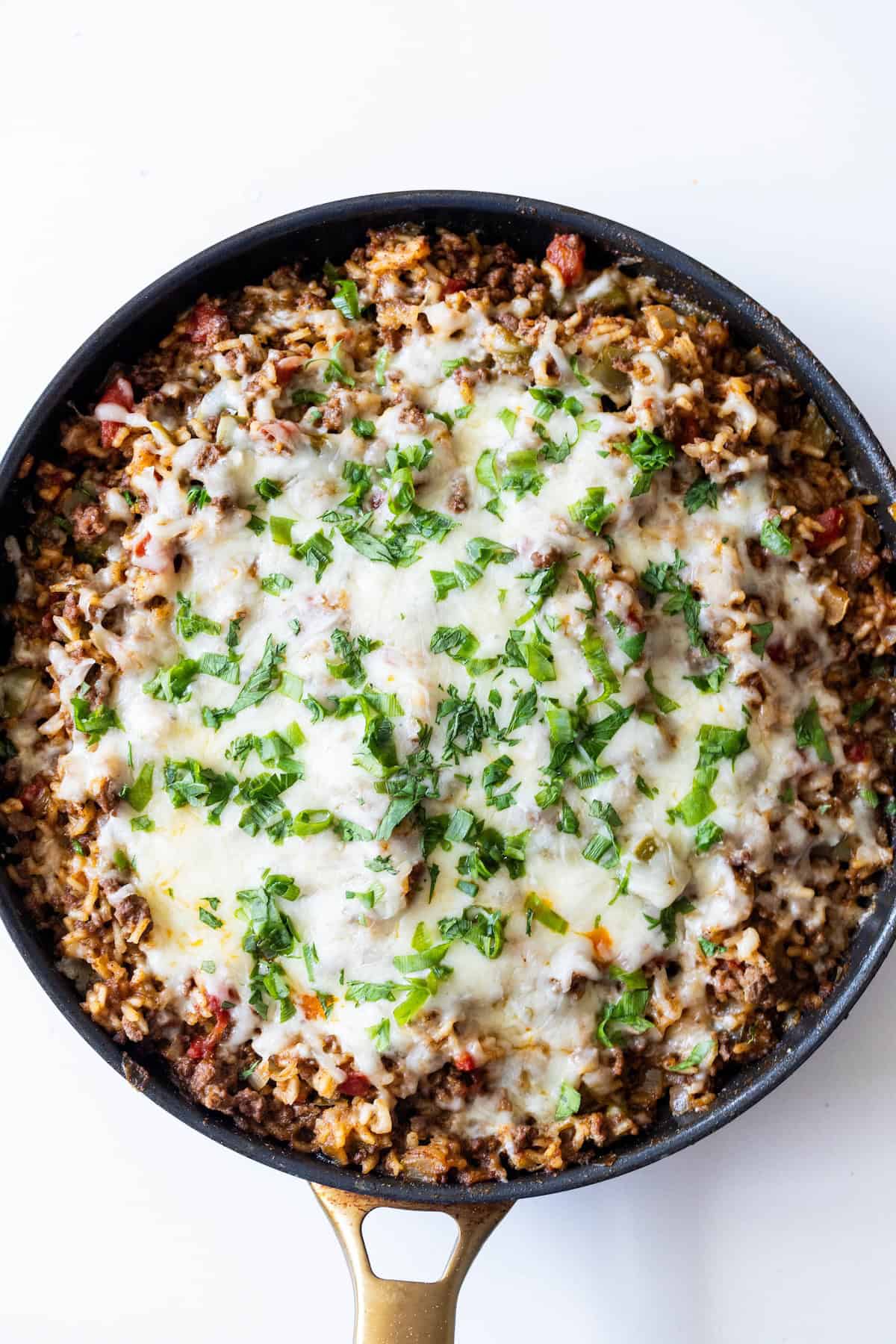 A skillet filled with ground meat, rice, tomato, melted cheese, and chopped green herbs on top—this One Pot Spicy Taco Rice Skillet is photographed from above.