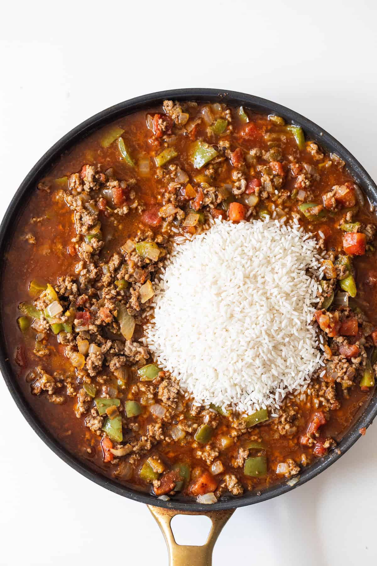 A skillet filled with ground beef, tomatoes, diced vegetables, and a mound of uncooked white rice on top—ready to be mixed for an easy taco and rice skillet dinner.