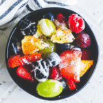 A black bowl filled with assorted fresh fruits such as grapes, strawberries, blackberries, blueberries, and pineapple, topped with a luscious lemon poppyseed icing for a delightful dessert fruit salad.