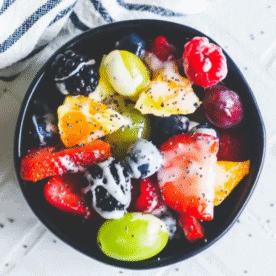 A black bowl filled with assorted fresh fruits such as grapes, strawberries, blackberries, blueberries, and pineapple, topped with a luscious lemon poppyseed icing for a delightful dessert fruit salad.