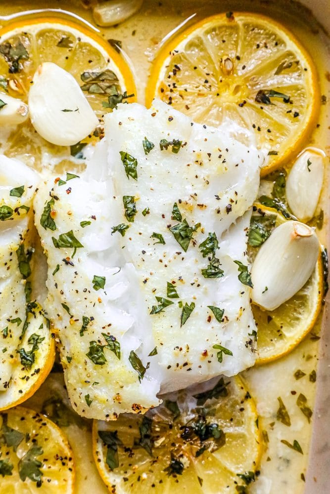 picture of baked cod in a ceramic dish with lemon slices and garlic cloves in the dish 