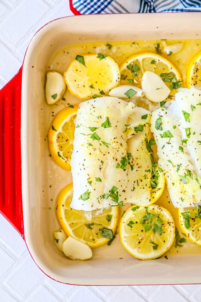 picture of baked cod in a ceramic dish with lemon slices and garlic cloves in the dish 