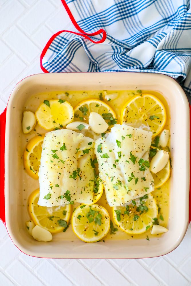 picture of baked cod in a ceramic dish with lemon slices and garlic cloves in the dish 