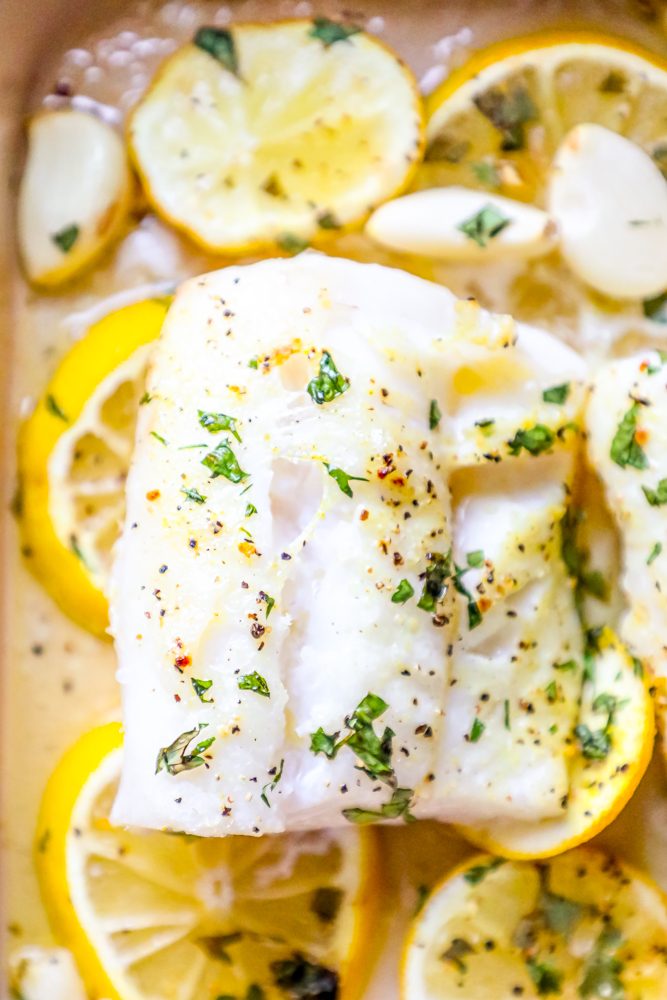 picture of baked cod in a ceramic dish with lemon slices and garlic cloves in the dish 