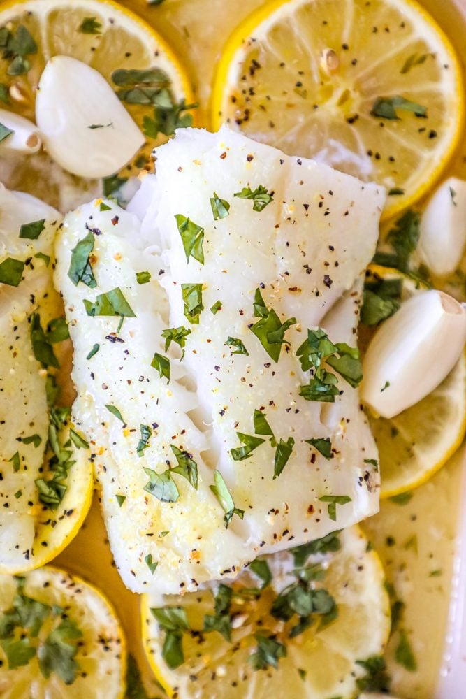 picture of baked cod in a ceramic dish with lemon slices and garlic cloves in the dish 