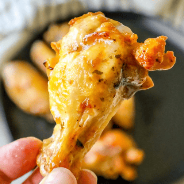 Close-up of a hand holding a cooked chicken drumstick with crispy, golden brown skin, resembling delicious keto ranch chicken wings, over a dark plate with more drumsticks.