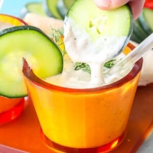 a slice of cucumber being dipped into tzatziki sauce in an orange cup