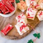 Fudge pieces with red candy chunks and Peppermint Crunch on a wooden board, next to a bowl of wrapped Andes mints and decorative string.