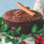 A Chocolate Chinese Five Spice Cake topped with cocoa powder, chocolate sprinkles, and two cinnamon sticks, decorated with green leaves and red berries.