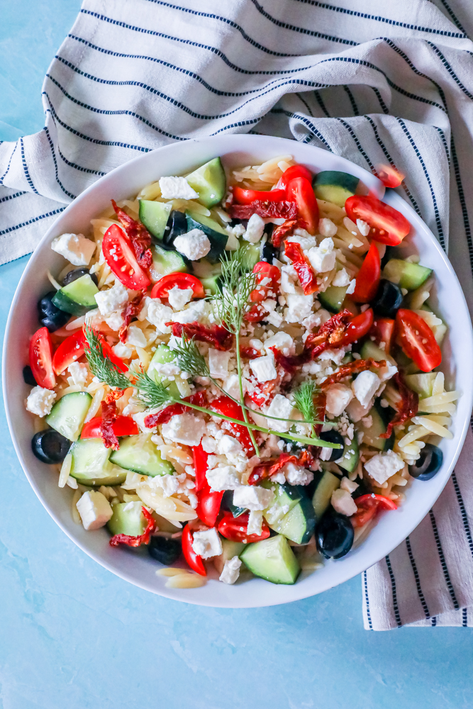cherry tomatoes, olives, sun dried tomatoes, feta cheese, cucumbers, and fresh dill in a bowl over orzo with a blue background.