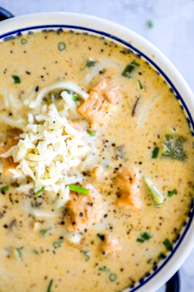 a spoon in a decorative blue bowl with creamy white chili, chunks of chicken, chopped bell peppers, and crumbled cheese on top in it