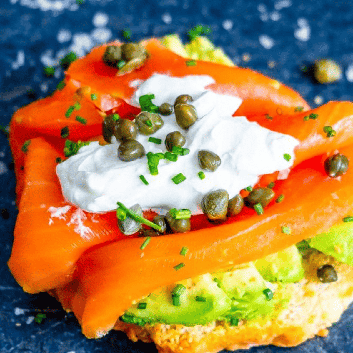 A close-up of a smoked salmon and avocado toast topped with cream cheese, capers, and chives on a blue plate.