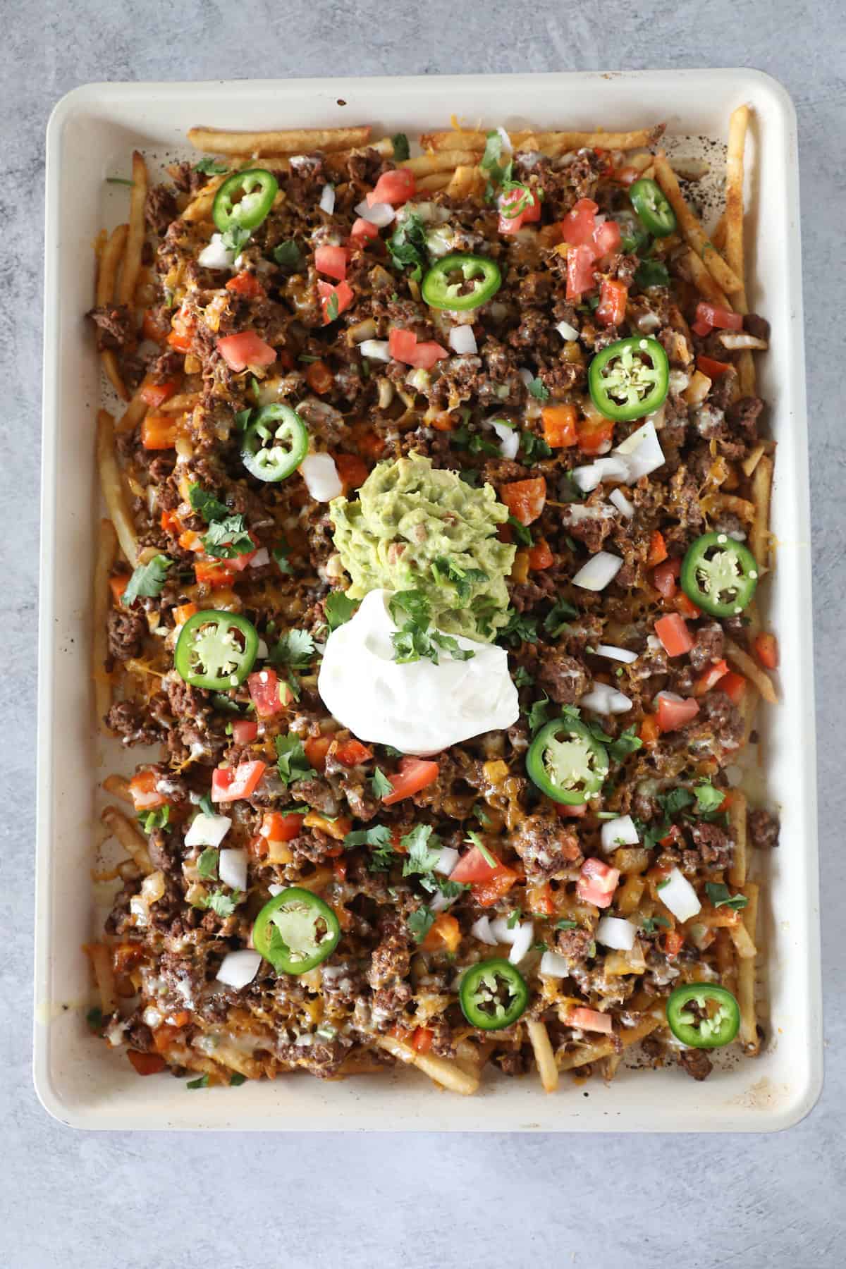 A baking sheet filled with loaded nacho fries topped with ground beef, melted cheese, diced tomatoes, onions, jalapeños, cilantro, guacamole, and sour cream for the ultimate snack.