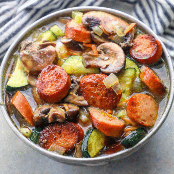 A bowl of keto sausage and vegetable soup recipe, featuring sliced sausage, mushrooms, zucchini, carrots, onions, and savory broth, sits on a gray surface with a striped cloth in the background.