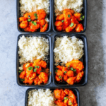 Four black plastic containers with sweet and sour chicken and rice in them.