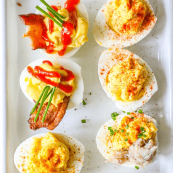 Six deviled eggs on a white tray, topped with bacon, chives, paprika, sriracha, and crab meat—these Easy Deviled Eggs Recipes are perfect for any gathering.