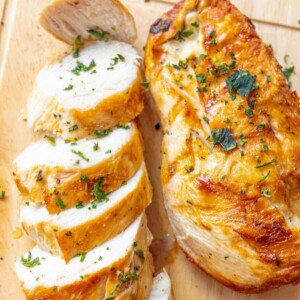 sliced grilled chicken breast next to grilled chicken breast with chopped parsley on a cutting board