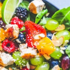 A colorful salad featuring fresh spinach, grapes, strawberries, blackberries, cubed chicken (the best chicken for fruit salad), sliced almonds, and lime wedges, garnished with sesame and poppy seeds. For extra zest, try a homemade citrus poppyseed dressing recipe.