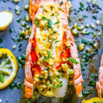 Cooked Lemon Garlic Butter Broiled Lobster Tails topped with herbs and butter, surrounded by lemon slices and chopped parsley on a baking sheet—a perfect low carb romantic dinner.