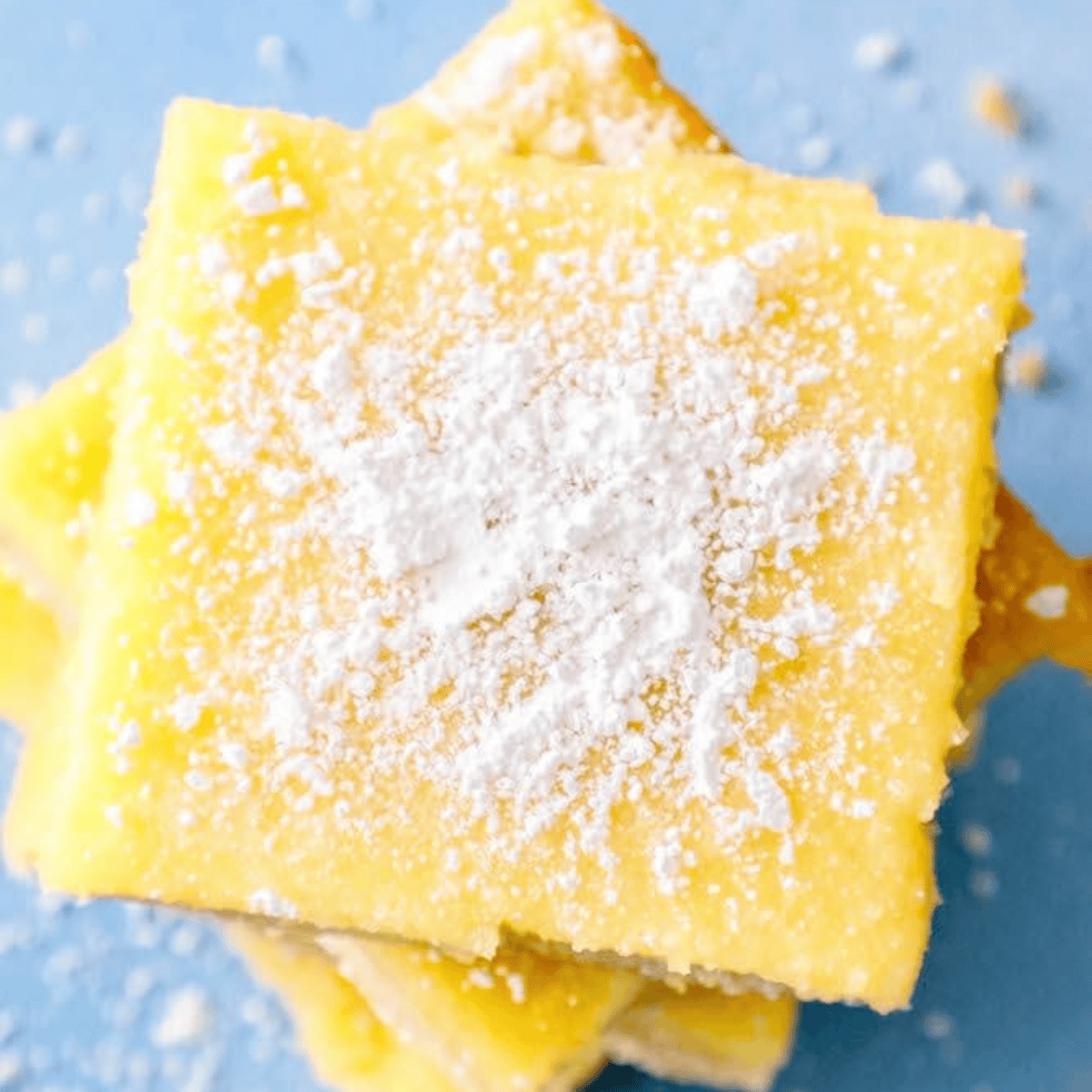 Close-up of the best lemon dessert bars stacked on a blue surface, delicately dusted with powdered sugar.