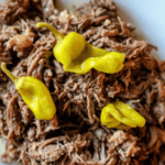 A plate of shredded meat from an Easy Mississippi Roast topped with three yellow chili peppers.