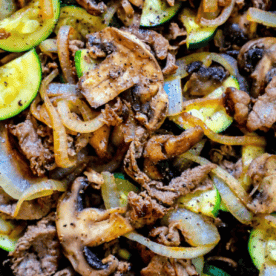 Close-up of a cooked dish featuring slices of beef, mushrooms, onions, and zucchini, seasoned with black pepper. This easy sirloin skillet with vegetables recipe is both savory and satisfying.