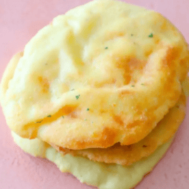 Two round, golden-brown, fluffy Keto Naan Bread pieces are stacked on a pink surface.