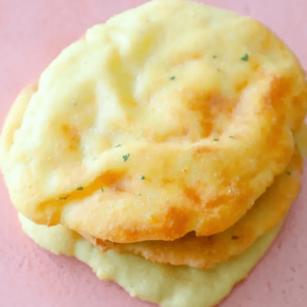 Two round, golden-brown, fluffy Keto Naan Bread pieces are stacked on a pink surface.