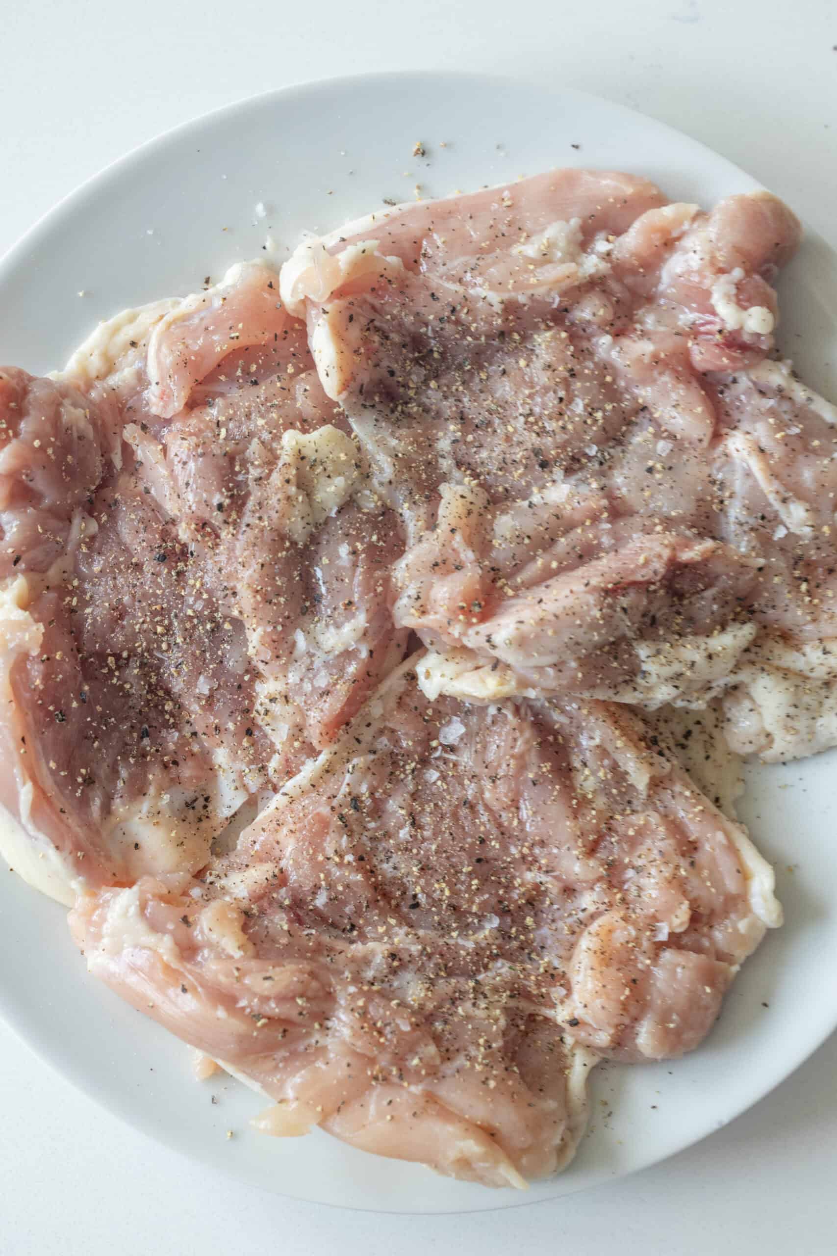 Raw chicken pieces seasoned with ground black pepper and salt on a white plate, perfect for making keto fried chicken thighs—no breading needed. Ideal for keto chicken thighs fried in butter for a flavorful, low-carb meal.