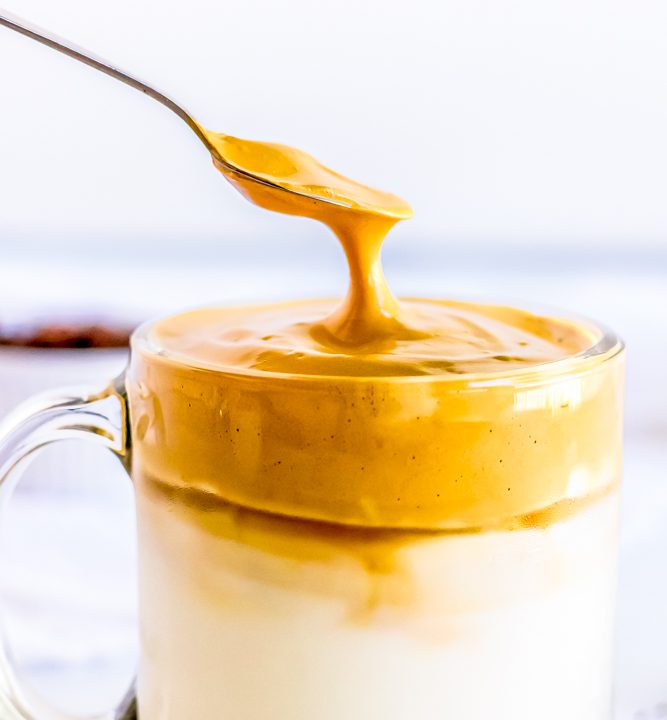 A spoon is being used to drizzle peanut butter on a cup of milk in an Easy Keto Dalgona Coffee Recipe.