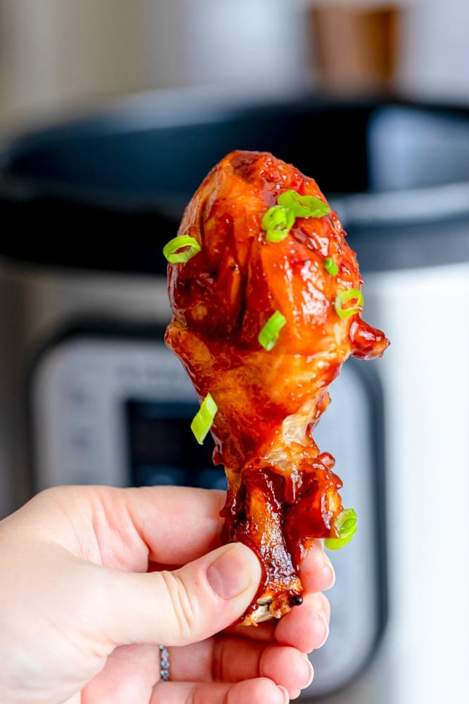 a hand holding a bbq chicken drumstick in front of an instant pot