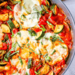 A close-up of an Easy Baked Zucchini Parmesan Skillet Recipe with melted cheese on top, featuring chunks of zucchini, red bell peppers, and garnished with fresh basil.